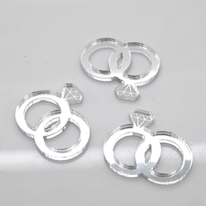 May include: Three silver mirrored acrylic wedding ring decorations. Each decoration features two interlocking rings with a diamond design.