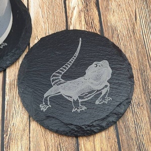 Peut inclure: Un dessous de verre en ardoise noire avec une image gravée en blanc d'un lézard dragon barbu.