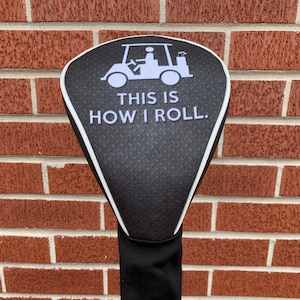 Puede incluir: Cubierta negra para la cabeza del palo de golf con un gráfico blanco de un carrito de golf y el texto "This is how I roll."