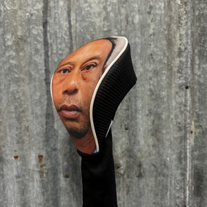 May include: A golf club head cover featuring a realistic image of a person's face. The cover has a black ribbed design on the back and a white trim. The bottom portion is black and cylindrical.