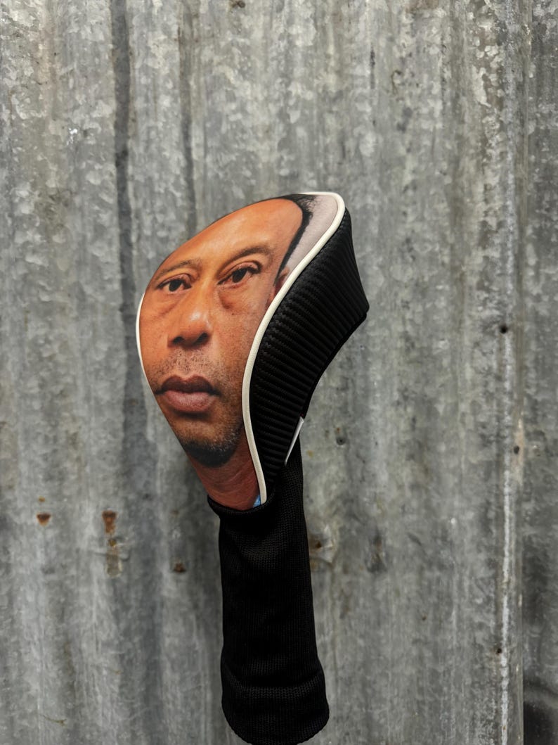 May include: A golf club head cover featuring a realistic image of a person's face. The cover has a black ribbed design on the back and a white trim. The bottom portion is black and cylindrical.