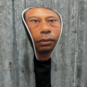 May include: A golf club head cover featuring a realistic image of a person's face. The cover has a black sock-like sleeve and a white trim around the face. The face is a medium skin tone.