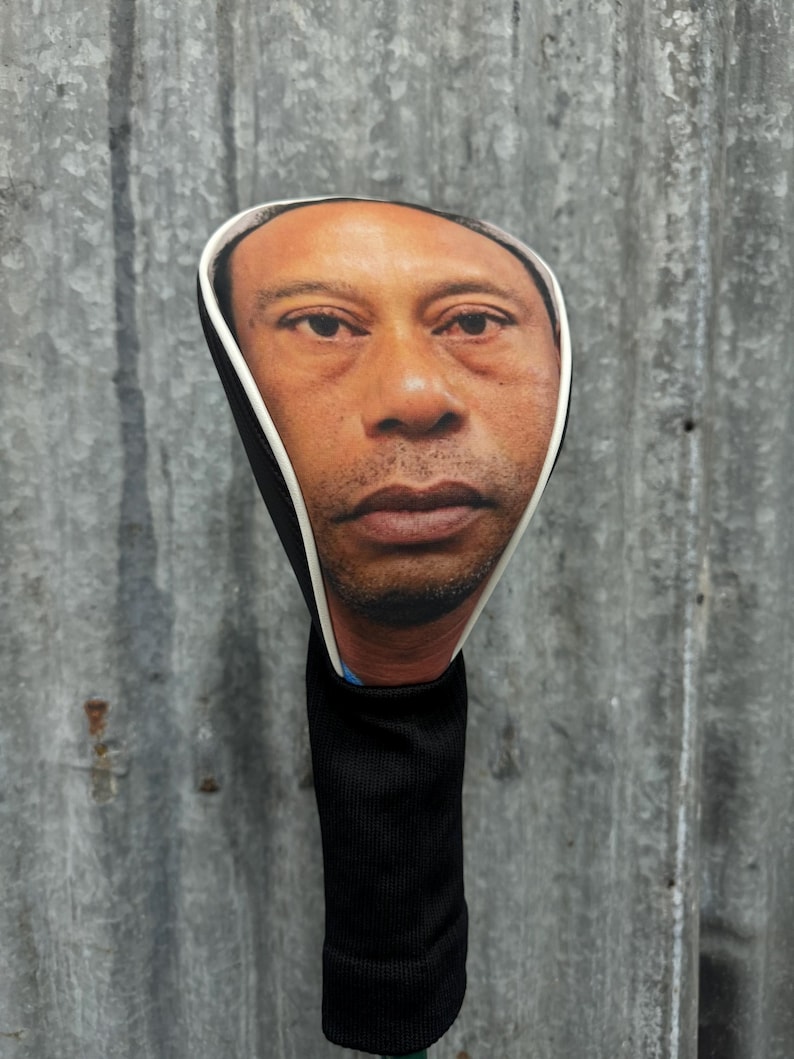 May include: A golf club head cover featuring a realistic image of a person's face. The cover has a black sock-like sleeve and a white trim around the face. The face is a medium skin tone.