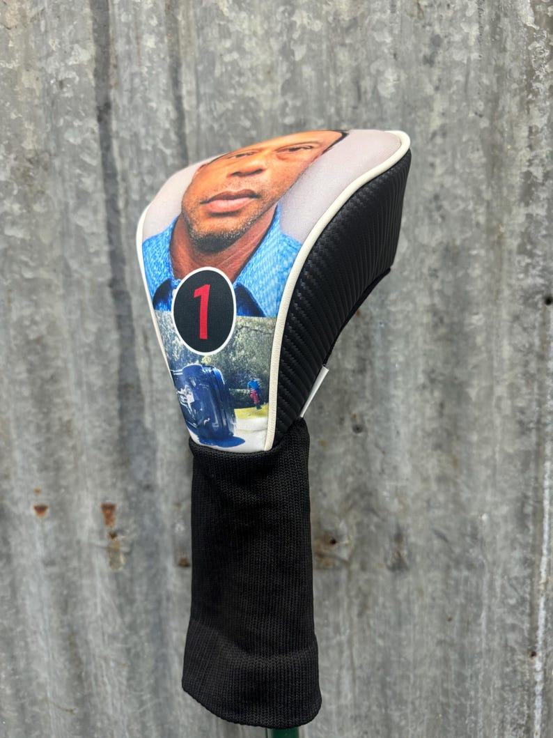 May include: Golf club head cover featuring a portrait of a man, with a black and white carbon fiber design on the sides. The cover has a black knit sock and a white trim. The number "1" is displayed on the front.