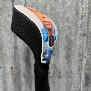 May include: A golf club head cover featuring a portrait of a person on the front. The cover has a black carbon fiber pattern on the sides and a black knit sock. The portrait is framed with a white border and the number 1 in red.