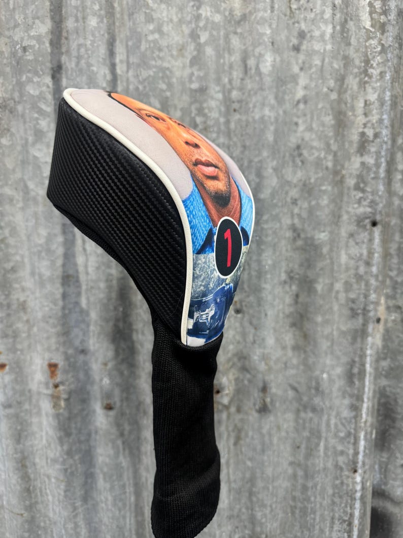 May include: A golf club head cover featuring a portrait of a person on the front. The cover has a black carbon fiber pattern on the sides and a black knit sock. The portrait is framed with a white border and the number 1 in red.