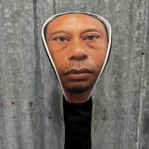 May include: A golf club head cover featuring a realistic image of a person's face. The cover has a black, ribbed sock-like body and a white-bordered face cutout. The background is a corrugated metal sheet.