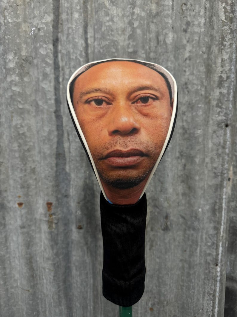 May include: A golf club head cover featuring a realistic image of a person's face. The cover has a black, ribbed sock-like body and a white-bordered face cutout. The background is a corrugated metal sheet.