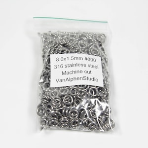 Może przedstawiać: Przezroczysta plastikowa torba wypełniona licznymi małymi, srebrnymi metalowymi pierścieniami. Biała etykieta na torbie głosi "8.0x1.5mm #800 316 stainless steel Machine cut VanAlphenStudio". Pierścienie są cięte maszynowo.