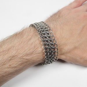 May include: A silver chainmail bracelet composed of multiple rows of small, interlocking metal rings. The bracelet is worn on a wrist, showcasing its intricate design and metallic sheen. The jewelry is a statement piece.