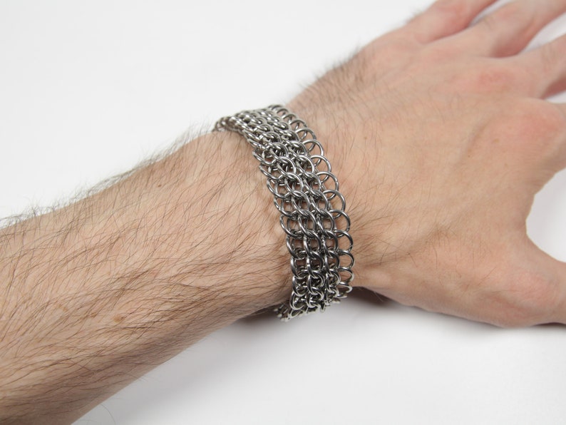 May include: A silver chainmail bracelet composed of multiple rows of small, interlocking metal rings. The bracelet is worn on a wrist, showcasing its intricate design and metallic sheen. The jewelry is a statement piece.