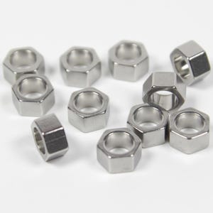 Könnte beinhalten: Eine Gruppe von silbernen sechseckigen Metallperlen mit einem Loch in der Mitte. Die Perlen sind auf einer weißen Oberfläche verstreut.