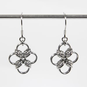 May include: A pair of silver earrings. Each earring has a four-petal design, with each petal formed by a circular loop. The loops are connected by woven metal, creating a textured, knotted appearance. The earrings hang from simple fish hook ear wires.