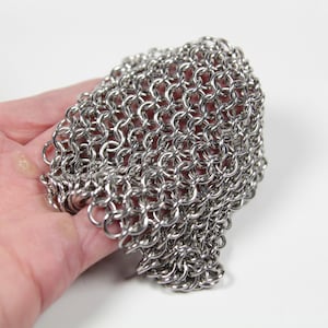 May include: Close-up of a stainless steel chainmail glove. The glove is made of small, interlocking metal rings, creating a flexible, protective mesh. The chainmail is held in a hand against a white background. This item is likely for historical reenactment or protection.