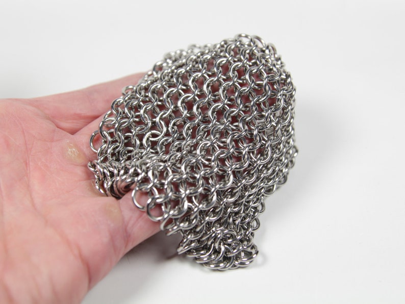 May include: Close-up of a stainless steel chainmail glove. The glove is made of small, interlocking metal rings, creating a flexible, protective mesh. The chainmail is held in a hand against a white background. This item is likely for historical reenactment or protection.
