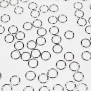 May include: A collection of small, shiny, silver-colored jump rings scattered on a white surface. These metal rings are circular and appear to be made of a durable material, suitable for jewelry making or crafting projects.