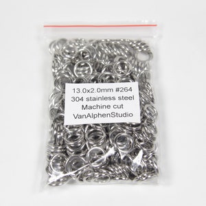 Może przedstawiać: Przezroczysta plastikowa torba wypełniona licznymi małymi, błyszczącymi, srebrnymi metalowymi pierścieniami. Torba ma białą etykietę z czarnym tekstem, który brzmi "13.0x2.0mm #264 304 stainless steel Machine cut VanAlphenStudio."