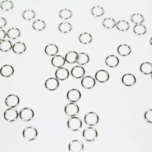 May include: A collection of silver jump rings, scattered on a white surface. The rings are various sizes and are used for jewellery making.