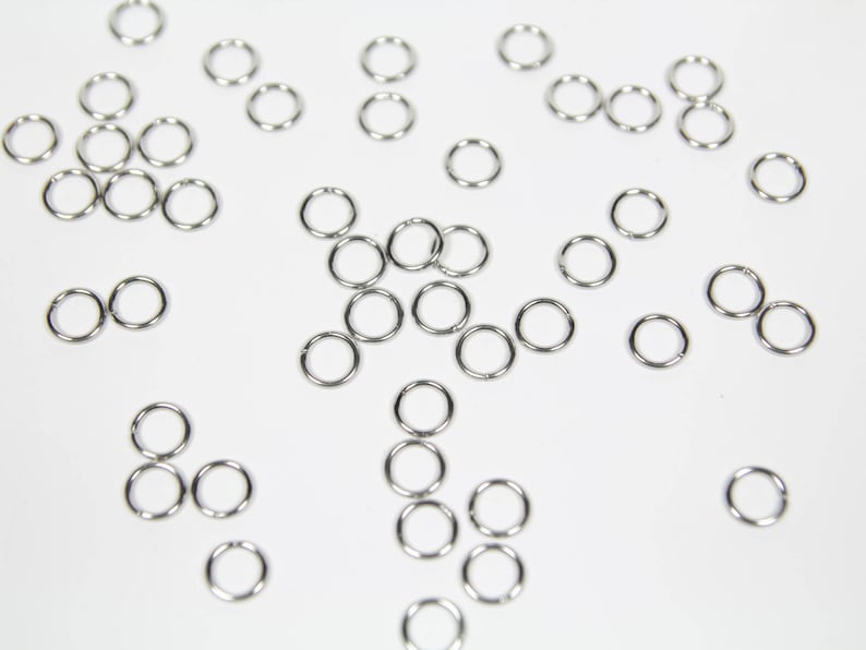 May include: A collection of silver jump rings, scattered on a white surface. The rings are various sizes and are used for jewellery making.