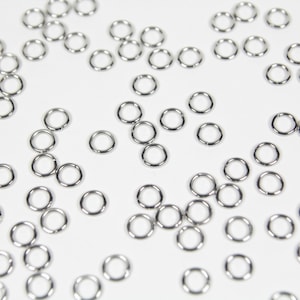May include: A collection of small, shiny, silver-colored jump rings scattered on a white surface. These circular rings are evenly spaced and appear to be made of metal, likely for jewelry making or crafts.