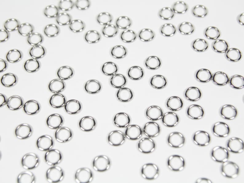 May include: A collection of small, shiny, silver-colored jump rings scattered on a white surface. These circular rings are evenly spaced and appear to be made of metal, likely for jewelry making or crafts.