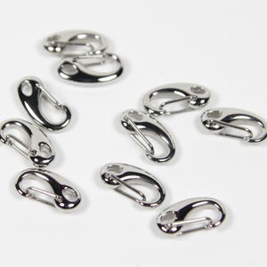 Könnte beinhalten: Eine Sammlung glänzender, silberfarbener ovaler Karabinerhaken. Diese Metallclips haben einen federbelasteten Verschluss für sicheres Befestigen. Die Clips sind auf einer weißen Oberfläche verstreut, ideal für verschiedene Bastel- und Hardware-Projekte.