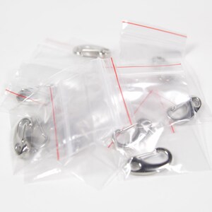 May include: A close-up of several small silver metal clasps in clear plastic bags. The bags have red lines along the edges.