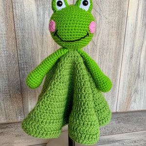 Puede incluir: Manta de beb&eacute; de ganchillo verde en forma de rana con mejillas rosas y ojos negros. La manta est&aacute; sobre un soporte plateado.