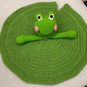 Puede incluir: Un nen&uacute;far de ganchillo verde con una rana de ganchillo en la parte superior. La rana tiene mejillas rosadas y ojos negros.