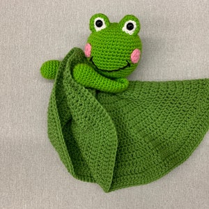 Puede incluir: Manta de seguridad de ganchillo verde en forma de hoja con una rana verde con mejillas rosas y ojos negros. La rana est&aacute; agarrando el borde de la manta.
