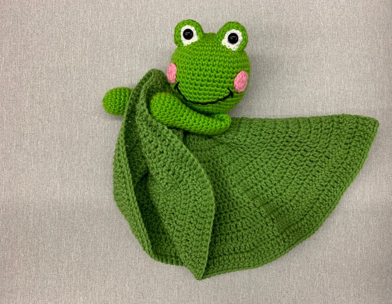 Puede incluir: Manta de seguridad de ganchillo verde en forma de hoja con una rana verde con mejillas rosas y ojos negros. La rana est&aacute; agarrando el borde de la manta.