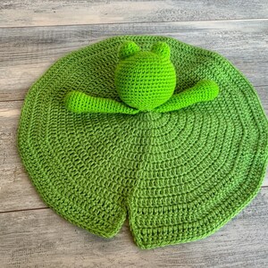Puede incluir: Un juguete de rana de crochet verde unido a un nen&uacute;far de crochet verde. El nen&uacute;far es de forma circular con una hendidura en el centro.