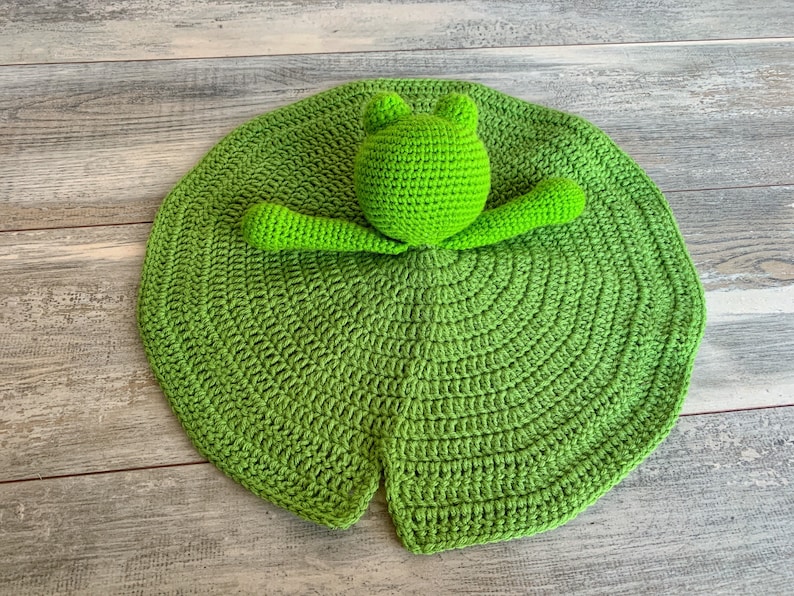 Puede incluir: Un juguete de rana de crochet verde unido a un nen&uacute;far de crochet verde. El nen&uacute;far es de forma circular con una hendidura en el centro.