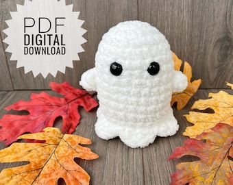 Puede incluir: Un fantasma blanco de crochet con ojos negros. El fantasma está sentado sobre una superficie de madera con hojas de otoño.