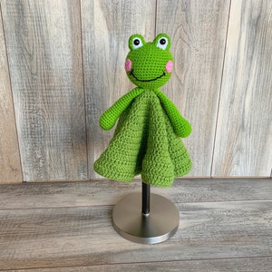 Puede incluir: Manta de beb&eacute; de ganchillo verde en forma de rana con mejillas rosas y una cara sonriente. La manta est&aacute; en un soporte.