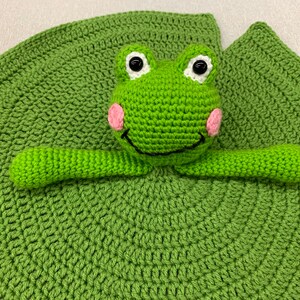 Puede incluir: Juguete de rana de crochet verde unido a un nen&uacute;far de crochet verde. La rana tiene ojos blancos, una sonrisa negra y mejillas rosadas.