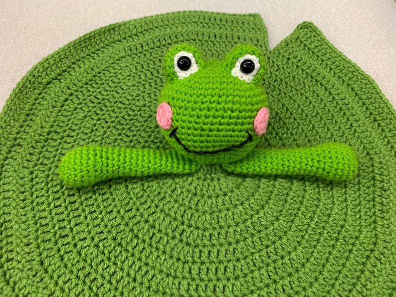 Puede incluir: Juguete de rana de crochet verde unido a un nen&uacute;far de crochet verde. La rana tiene ojos blancos, una sonrisa negra y mejillas rosadas.