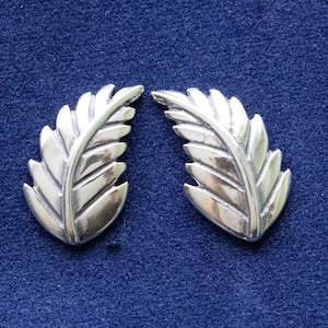 May include: Two silver fern leaf earrings, each measuring approximately 1 inch in length. The earrings are shown against a blue background with a ruler for scale.