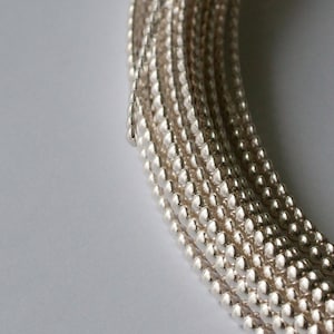 Könnte beinhalten: Nahaufnahme einer silbernen Kette aus kleinen, runden Perlen. Die Kette ist aufgerollt und die Perlen sind dicht aneinander gereiht.