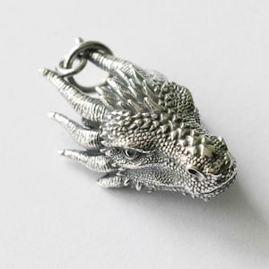 May include: A silver dragon head charm with detailed scales and claws. The charm has a loop for attaching to a necklace or bracelet.