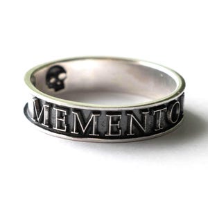 May include: A silver ring featuring the word "MEMENTO" engraved around the band. A small skull is visible on the ring's surface. The ring is set against a white background.