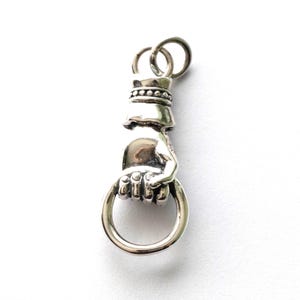 May include: A silver pendant depicting a hand grasping a ring. The hand is intricately detailed with sculpted fingers and a textured wrist. A small crown design sits above the hand, with a loop for a chain.