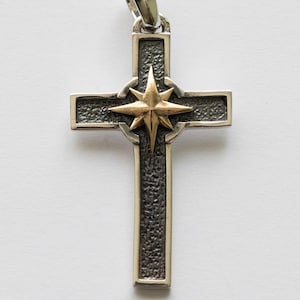 May include: Silver-tone cross pendant with a textured surface and a gold-colored starburst design at the center. The pendant has a bail for attaching to a chain. A religious symbol.