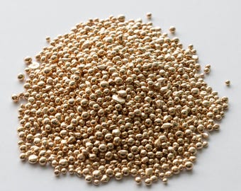 May include: A close-up of a pile of small, round, gold-colored metal beads. The beads are scattered on a white surface.