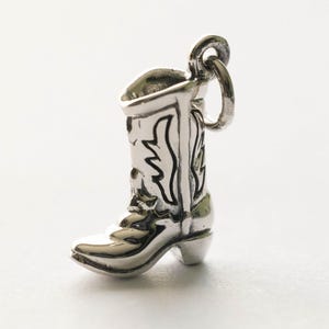 May include: A silver charm in the form of a cowboy boot. The boot features etched patterns and a loop for attachment. This miniature charm is a detailed representation of a traditional Western boot, suitable for a bracelet or necklace.
