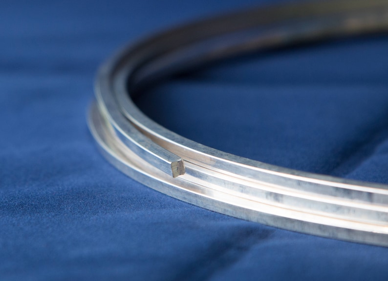 Puede incluir: Primer plano de tres tiras de metal plateado, cada una con una secci&oacute;n transversal cuadrada, apiladas una encima de la otra. Las tiras est&aacute;n sobre un fondo de tela azul.