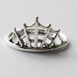 以下が含まれることがあります： クモの巣のデザインが施されたシルバーカラーのリング。リングは丸みを帯びたバンドと、上部をアーチ状に横切るウェブのような構造を備え、ユニークでゴシックな美しさを生み出しています。リングはおそらくスターリングシルバー製です。