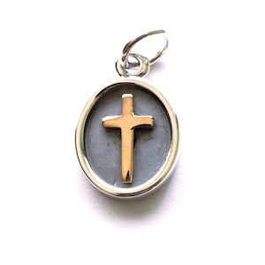 May include: A silver pendant with a gold cross set against a gray background. The cross is centered within an oval frame, and the pendant has a small loop at the top for a chain.