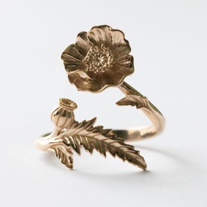 May include: A gold-colored ring featuring a detailed poppy flower design. The ring's band curves around the finger, with a poppy flower and a seed pod accent. The ring is made of a shiny, metallic material, and the design is intricate.