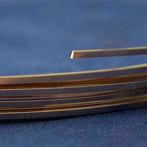 以下が含まれることがあります： ジュエリー作りに使用される可能性のある、金色のワイヤーの束のクローズアップ。ワイヤーは平らで断面が長方形で、明るく反射する表面を持っています。背景は無地の濃い青色です。